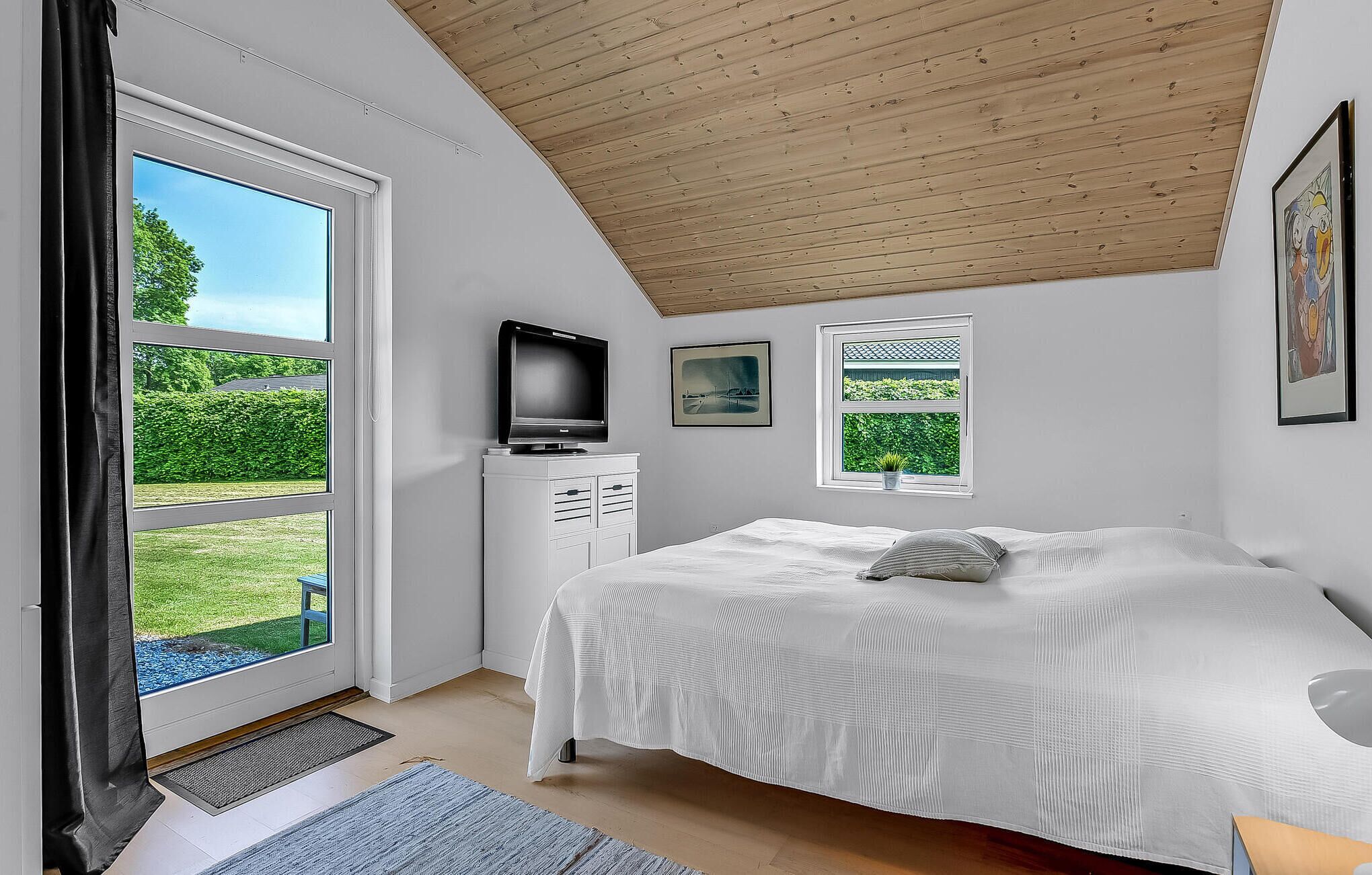 Une chambre avec une grande fenêtre, un lit, une télévision et un petit meuble.