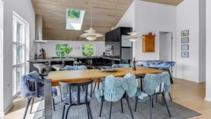 Una cocina moderna con techo de madera, un comedor con mesa y sillas de madera, y vistas al exterior a través de grandes ventanales.
