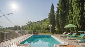 Une piscine rectangulaire, bordée d’une terrasse carrelée et entourée d’une clôture en métal. On y trouve des chaises longues et un parasol vert sur le côté droit.