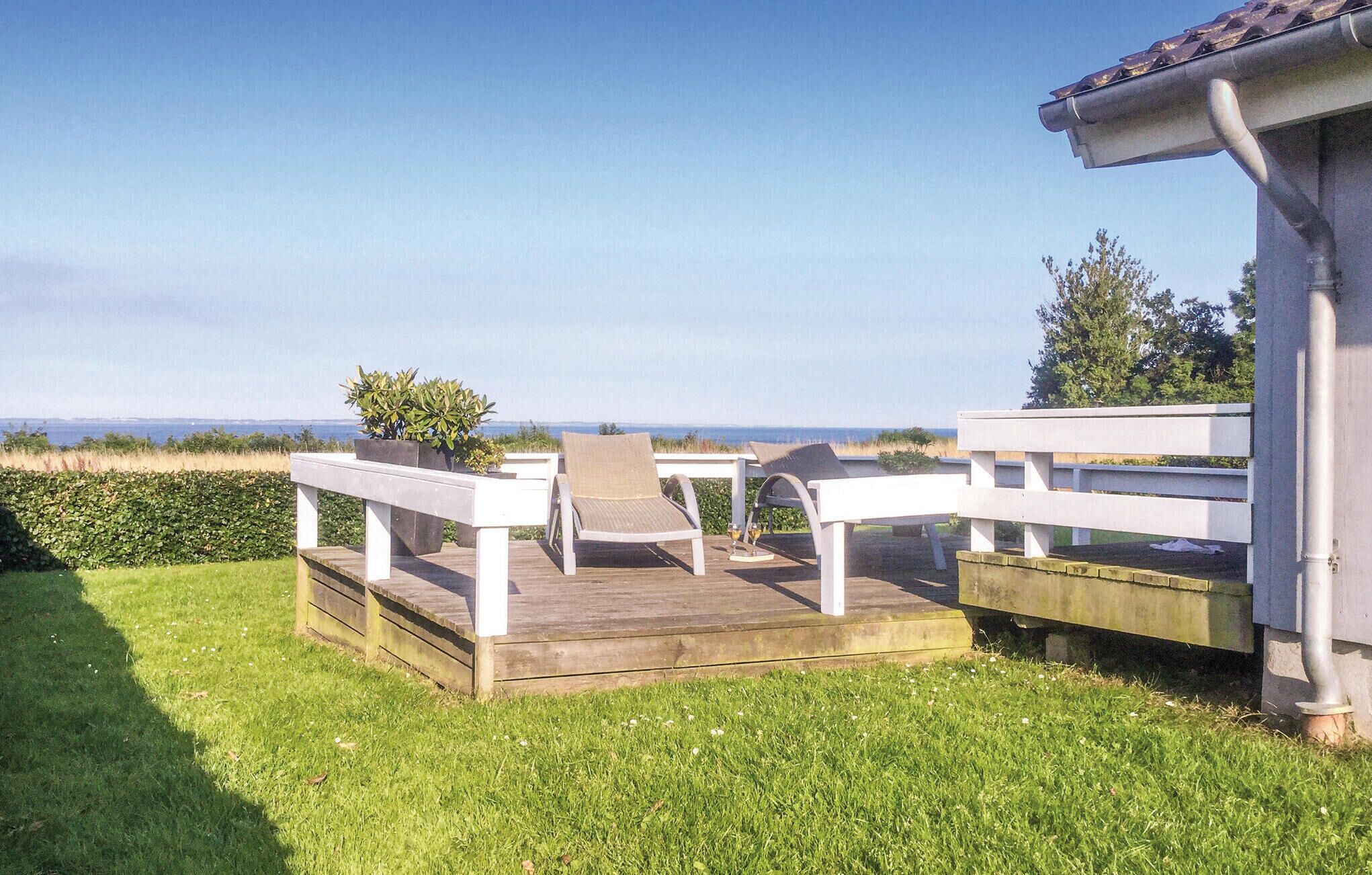 A wooden deck with white benches and chairs, overlooking a grassy area with a view of the sea.