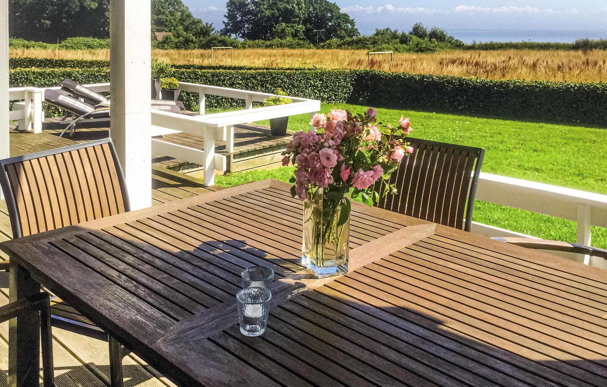 Une table en bois sur laquelle sont posés un vase rempli de fleurs roses et un verre d’eau, le tout installé sur une terrasse dotée de rampes blanches et offrant une vue sur un champ.