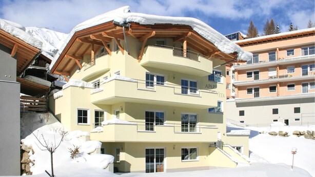 Un edifici d'alguns pisos amb balcons i un teulat cobert de neu, envoltat d'altres edificis.