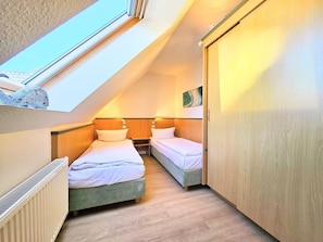 A small, cozy attic bedroom with two single beds, a wooden wardrobe, and a skylight.