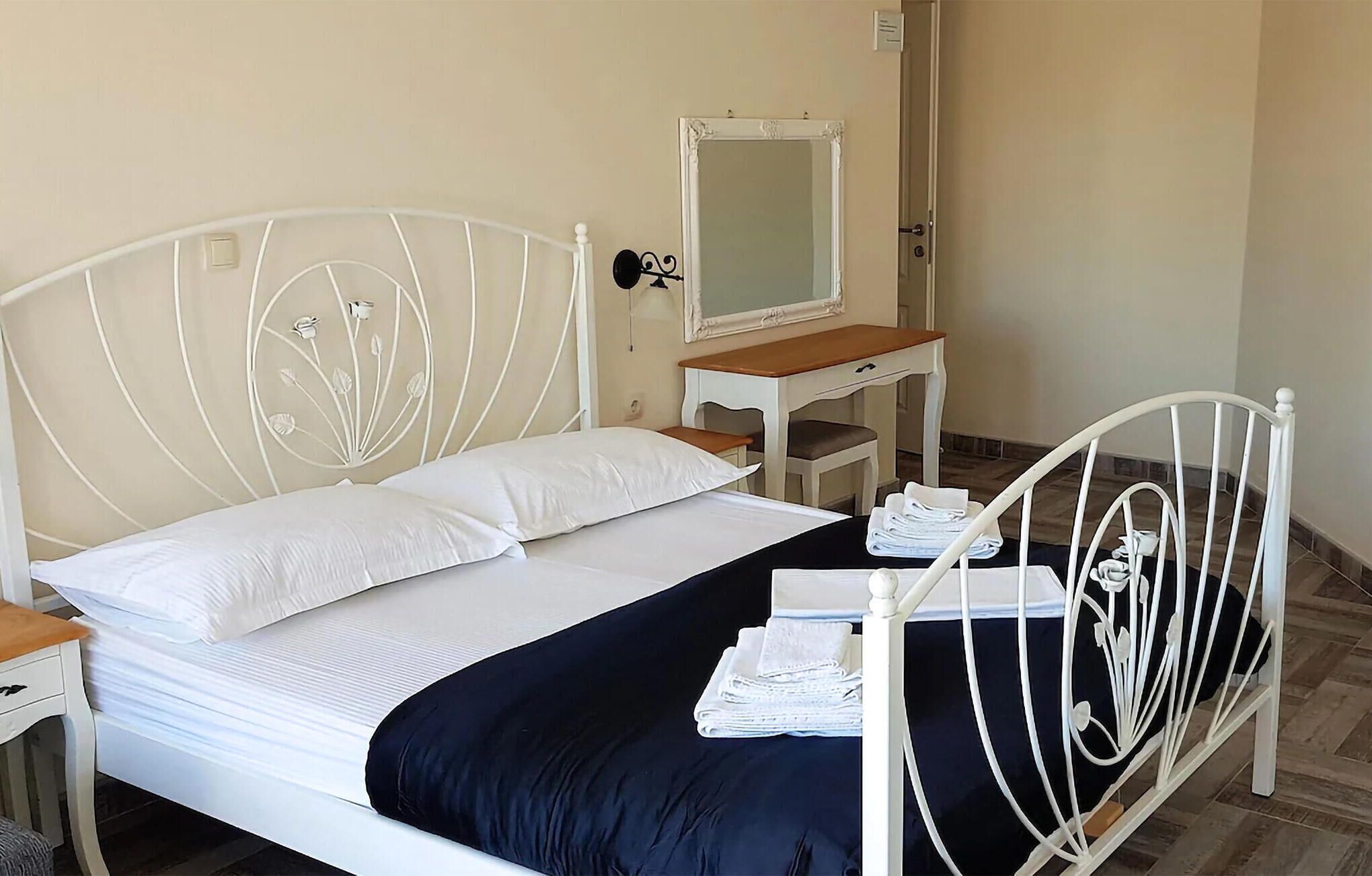 Un lit bien fait, avec du linge de lit blanc et une couvre-lit bleu foncé, une petite table de chevet avec une lampe, une table de nuit en bois et un miroir au mur.