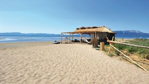 A beach with a wooden structure and a thatched-roof hut, a boat, and a clear view of the sea and distant mountains.