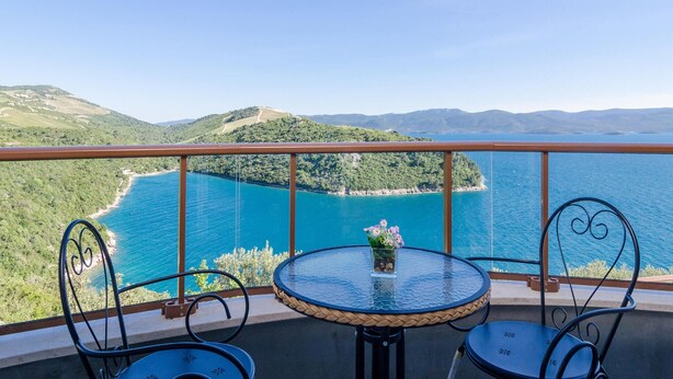 A balcony with a glass table and two chairs, overlooking a turquoise sea and green hills.