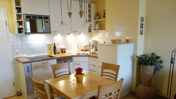 A compact kitchen with white cabinets, a wooden dining table, and chairs. There is a sink, a coffee maker, and a potted plant. - (191) 2-room apartment - apartment house Baltic Sea beach (Kühlungsborn)