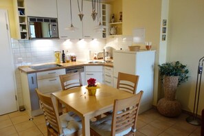 Una cucina compatta con mobili bianchi, un tavolo da pranzo in legno e sedie. Si vede un lavandino, una moka e una pianta in vaso.