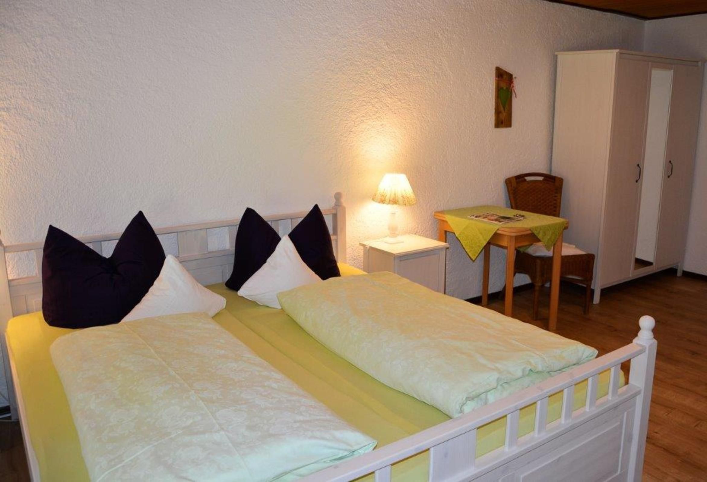 Ein Doppelbett mit Kissen, ein Nachttisch mit Lampe, ein kleiner Tisch mit gelber Tischdecke und ein Kleiderschrank.