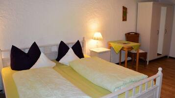 A double bed with pillows, a bedside table with a lamp, a small table with a yellow tablecloth, and a wardrobe.