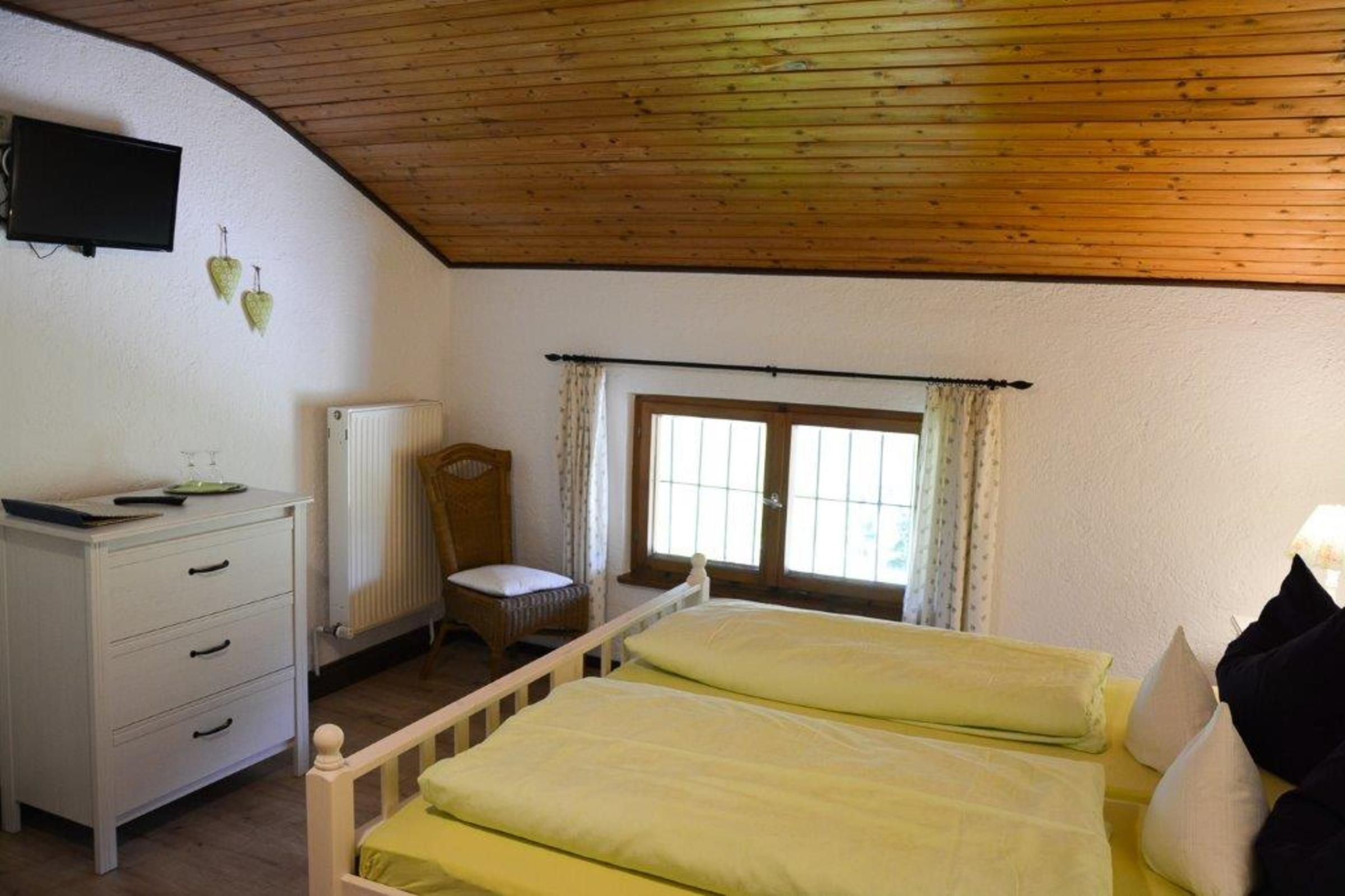 Ein Schlafzimmer mit Holzbalkendecke, einem Bett mit gelber Bettwäsche, einem weißen Schminktisch, einem Sessel, einem Heizkörper und einem Fenster mit Vorhängen.