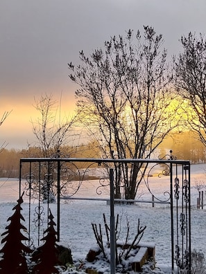A metal gate with intricate designs, a snow-covered landscape, and leafless trees against a sunset. - Double room Hollerbeerl - Hof Roßruck (Fischbachau)