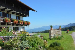 A traditional chalet with a balcony adorned with flowers, a stone wall, and a view of mountains. - Double room Hollerbeerl - Hof Roßruck (Fischbachau)
