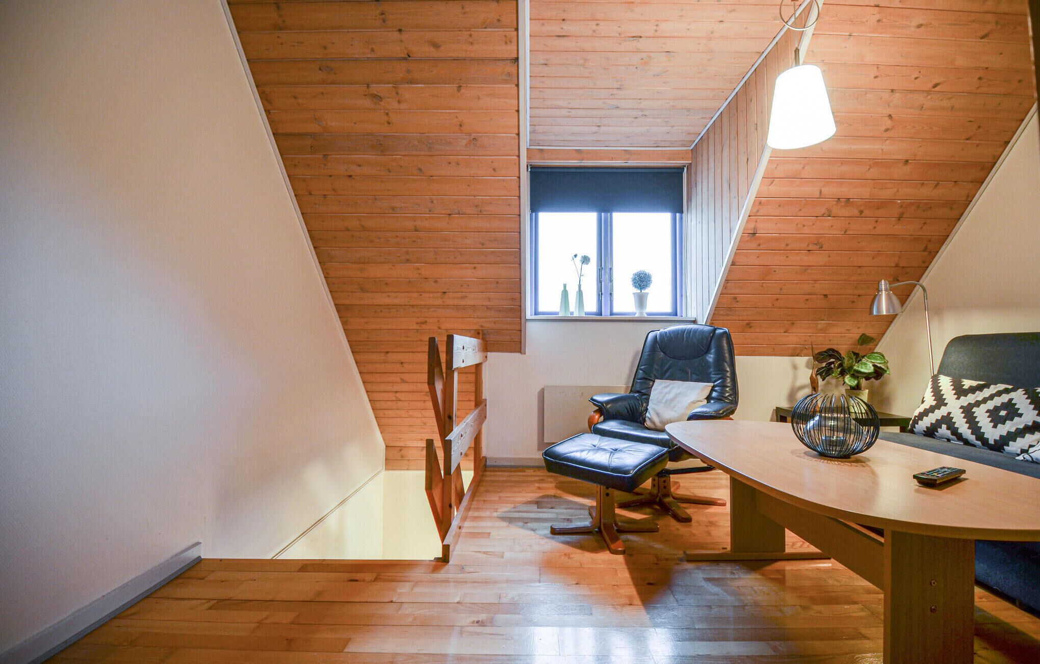 A cozy attic room with wooden floors, a black leather chair, a wooden table, and a window with a view.