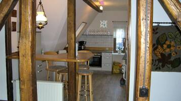 A rustic kitchen with wooden beams, a dining table with chairs, a hanging light fixture, and a view of the living area.