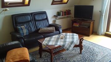 A living room with a black leather sofa, a small round table, a television on a wooden stand, and a patterned rug.