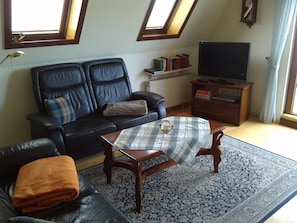 A living room with a black leather sofa, a small round table, a television on a wooden stand, and a patterned rug. - Vacation apartment Villa am Wald (Blankenburg (Harz))
