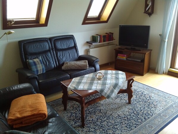 Ein Wohnzimmer mit einer schwarzen Ledersofa, einem kleinen runden Tisch, einem Fernseher auf einem Holzständer und einem gemusterten Teppich.