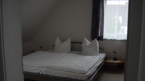 Ein Einzelbett in einem Zimmer mit schrägen Wänden, einem kleinen Nachttisch und einem fenster mit Vorhängen.