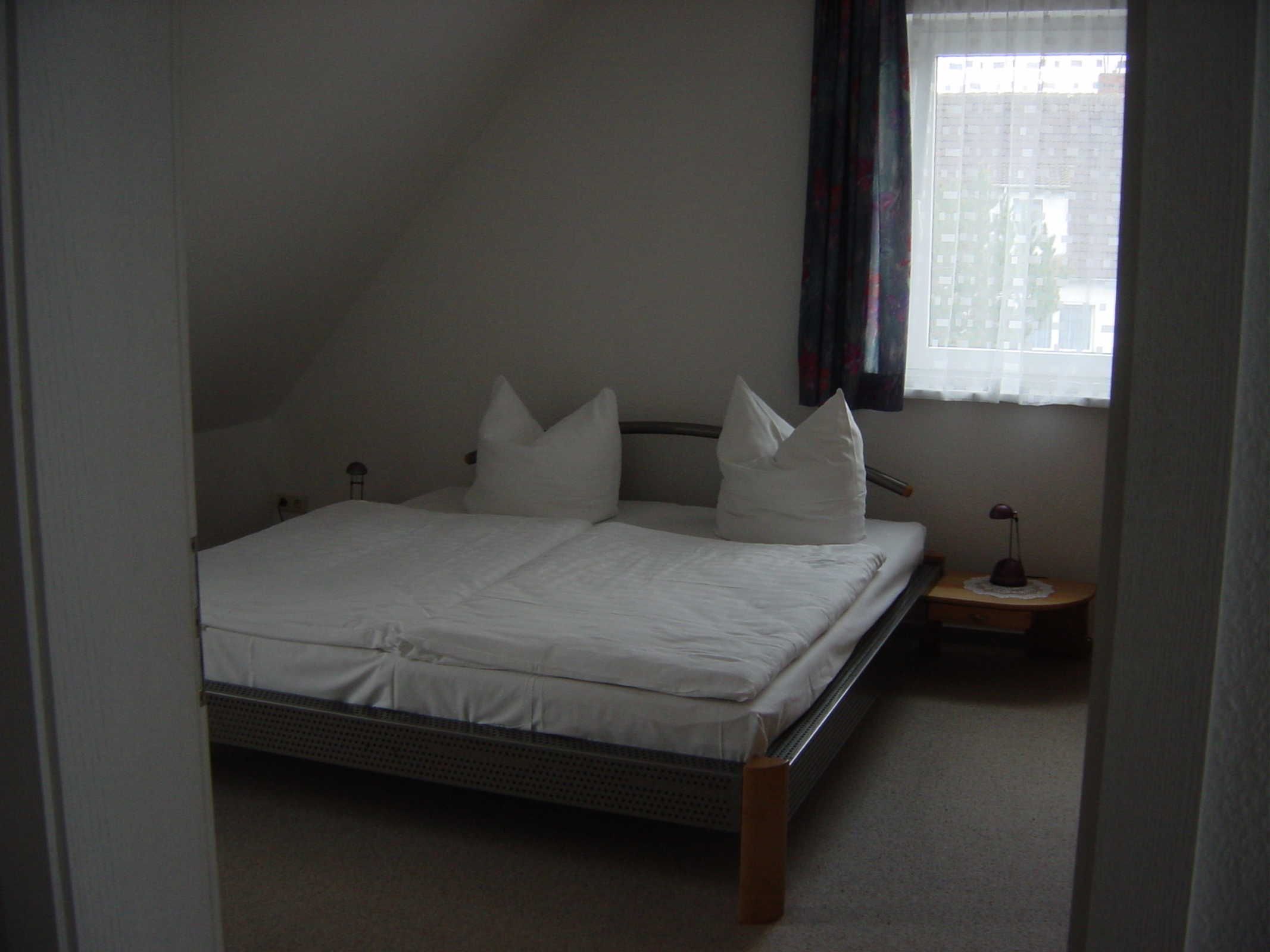 A single bed in a room with a sloped ceiling, a small bedside table, and a window with curtains.