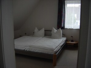 A single bed in a room with a sloped ceiling, a small bedside table, and a window with curtains. - Apartment 1 floor - house gallery (Karlshagen)