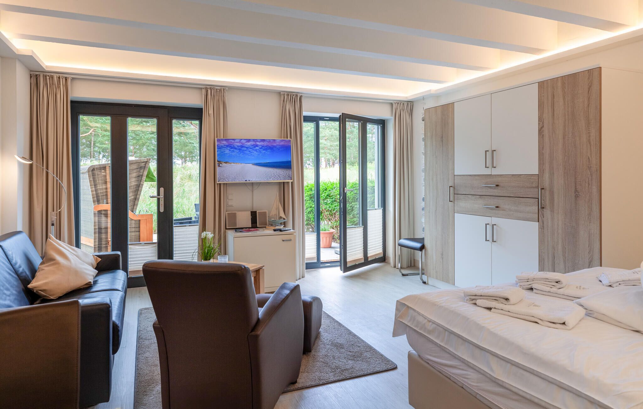 A modern hotel room with a sofa, a bed, a TV, and a balcony with a view of greenery.