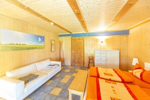 A room with a wooden ceiling, a white sofa, a bed with orange bedding, a wooden table, and a painting on the wall.