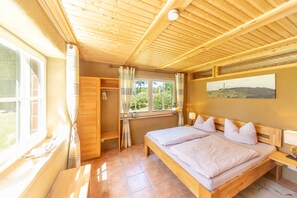 A bedroom with a wooden ceiling, a large bed, a window with curtains, a painting on the wall, and a wooden bench by the window. - Holiday EC - Apartments in thatched house (Ummanz)