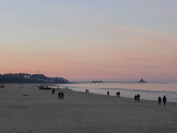A beach with a pier extending into the water, people walking along the shore, and buildings in the background.