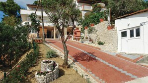 A residential building with a paved pathway leading to a door, surrounded by greenery and a stone wall.