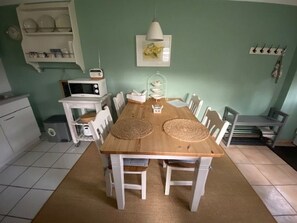 Uma sala de jantar com mesa de madeira, cadeiras brancas, armário branco, micro-ondas e luminária pendente.