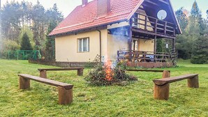 Une maison à deux étages avec un toit rouge et un balcon. Un feu brûle devant la maison.