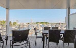 Een overdekt terras met tafels en stoelen, met uitzicht op een jachthaven vol boten en een verre skyline.