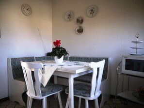 A dining area with a white table, two chairs, a vase with red flowers, a clock, and decorative plates on the wall.