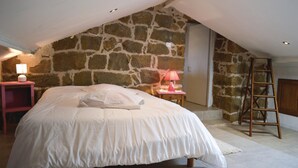 Un dormitorio con pared de piedra, cama con ropa blanca, lámpara rosa sobre una mesita de noche y una escalera de madera.