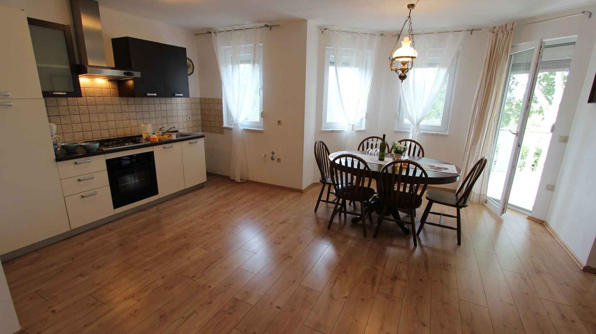 A kitchen with wooden flooring, a dining table with chairs, a chandelier, and a view of the outdoors through windows.