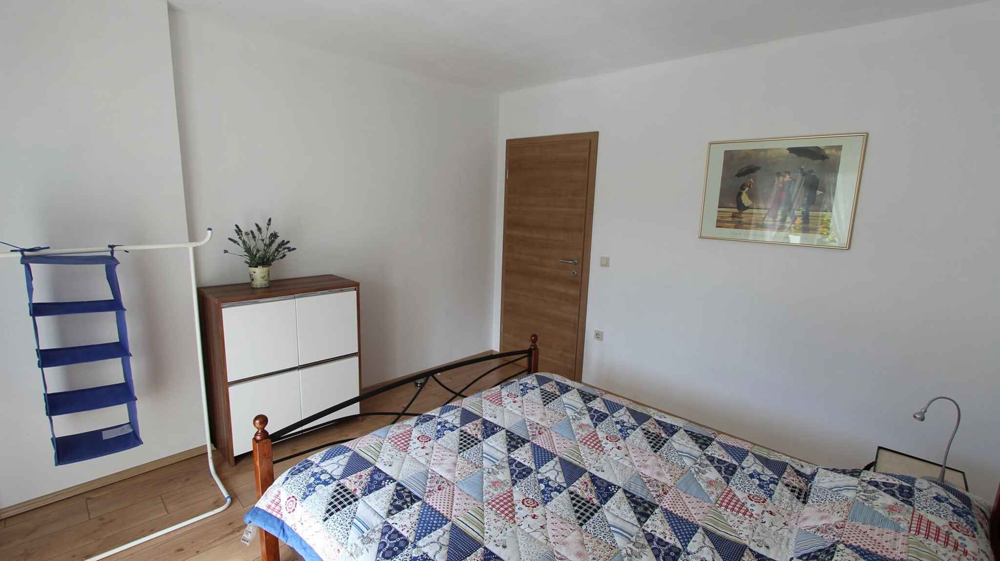 Un dormitorio con cama, un armario de madera, una puerta, un cuadro enmarcado en la pared y una planta sobre el armario.