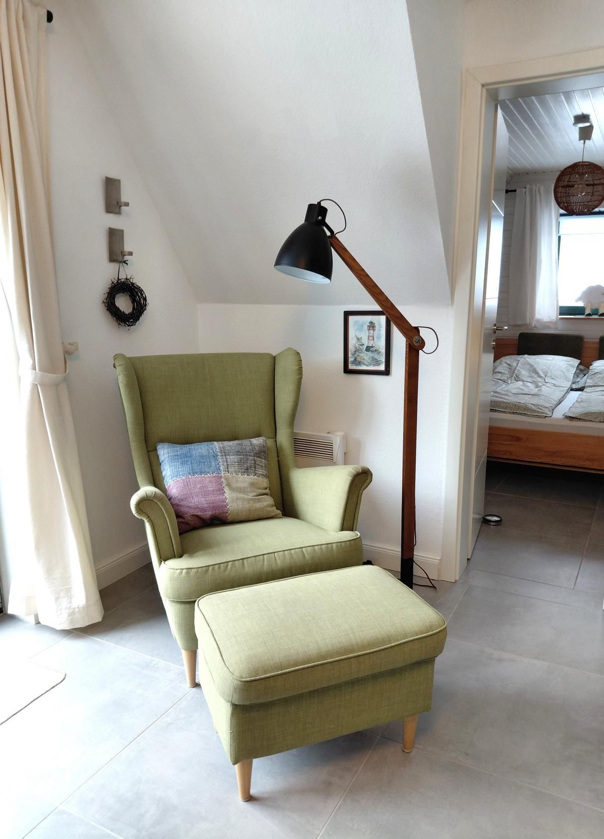 A room with a green armchair, a small ottoman, a floor lamp, a framed picture, and a bed in the background.