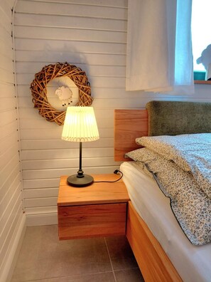 Un dormitorio con mesita de noche de madera, un arreglo de mimbre en la pared, una lámpara de noche y una cama con ropa de cama estampada con motivos florales.