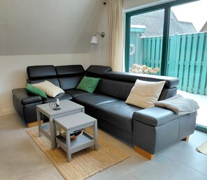Un salon moderne avec un canapé d’angle, une petite table d’appoint et une grande fenêtre donnant sur une clôture verdoyante.