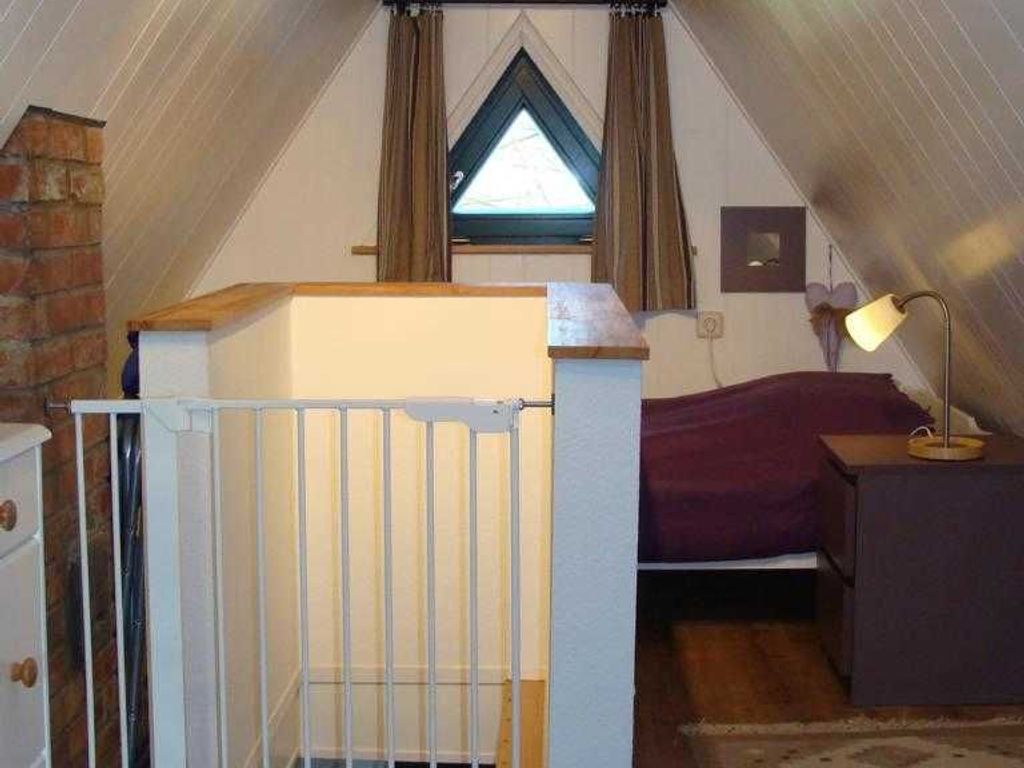 Ein kleines Schlafzimmer unter der Dachschräge, mit einem Fenster mit Vorhängen, einem Bett, einem Nachttisch und einem weißen Heizkörper.