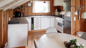 Een kleine keuken met witte kasten, een roestvrijstalen koelkast en een inbouwoven. Boven de gootsteen is een raam en er hangt een klok aan de muur.