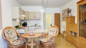 A compact kitchen with white cabinets, a dining area with wicker chairs, and a wooden cabinet with a glass door. - (170) 1-room apartment - apartment house Sanssouci (Kühlungsborn)