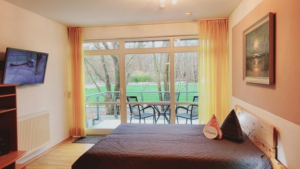 A bedroom with a bed, a television, a window with curtains, and a view of the outdoors. - (170) 1-room apartment - apartment house Sanssouci (Kühlungsborn)