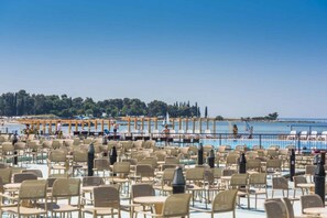Un espace aménagé au bord de la piscine, avec de nombreuses chaises et tables, une vue dégagée sur la mer et, au loin, une côte bordée d’arbres et de bâtiments.