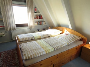 A bedroom with a sloped ceiling, two single beds with striped bedding, a wooden nightstand, a bookshelf with books, a radiator, and a window with curtains.