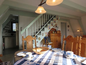 A dining area with a checkered tablecloth, wooden chairs, a basket of bread, and a staircase in the background.