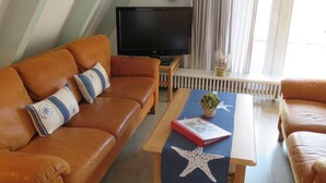 Ett vardagsrum med en brun lädersoffa, ett träbord med en sjöstjärna som dekoration, en röd bok och en liten krukväxt. En tv är monterad på väggen ovanför soffan.