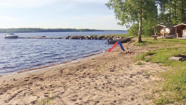 A sandy beach with a boat docked on the shore, a pier extending into the water, and a row of cabins in the background. - Amazing home in Bolmsö with sauna (Bolmsö)