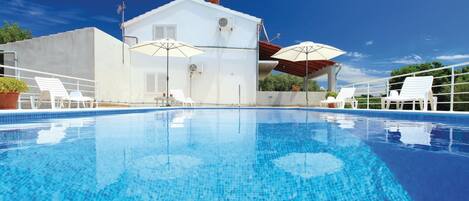 A clear blue pool with white lounge chairs and umbrellas, a white building with a red roof, and a clear sky.